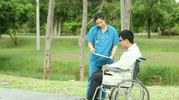 Lycklig asiatisk man Sammanträde på rullstol i de parkera och ta vård förbi asiatisk sjuksköterska på helger Semester video