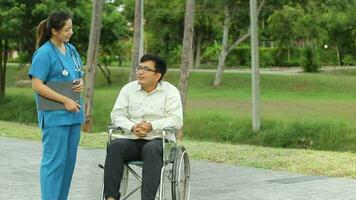 Happy Asian man sitting on wheelchair in the park and take care by asian nurse on weekends holiday video