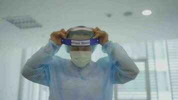 Doctor man in the protective mask is putting on a face shield and looking at you while standing in the cabinet of a modern clinic video