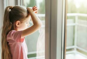 triste pequeño niña mirando mediante ventana a hogar. foto