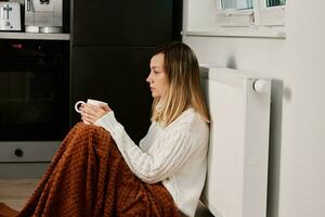 Worried woman sit near heating radiator under blanket with cup of tea photo