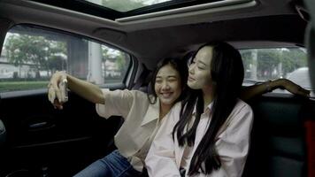 Beautiful young girls taking selfie with their friend on phone or smartphone in back seat of moving car while roadtrip. Cheerful friends having good time together. Concept of joy and lifestyle video