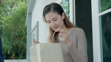 une femme asiatique souriante et heureuse reçoit un colis de nourriture en sac en papier de la maison de messagerie. livreur envoyer livrer express. achats en ligne, conteneur en papier, plats à emporter, facteur, service de livraison, colis video