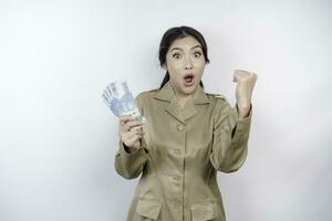 A successful young government worker woman is wearing khaki uniform holding cash money in Indonesian rupiah isolated by white background photo