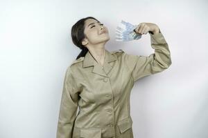 A happy young government worker woman is wearing khaki uniform holding cash money in Indonesian rupiah isolated by white background photo