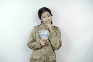 A thoughtful young government worker woman is wearing khaki uniform holding cash money in Indonesian rupiah isolated by white background photo