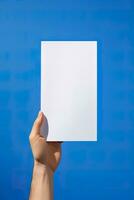 A human hand holding a blank sheet of white paper or card isolated on blue background. ai generated photo