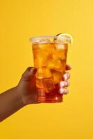 A hand holding takeaway plastic cup of delicious iced lemon tea isolated on yellow background. ai generated photo