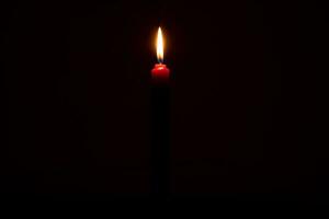 Single burning candle flame or light glowing on a small red candle on black or dark background on table in church for Christmas, funeral or memorial service with copy space. photo