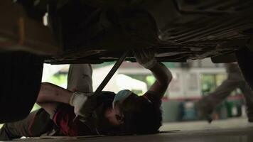 Asian man mechanic lying under car using wrench tightening nut repairing undercarriage of car in workshop. Repair and maintenance of the car in service. video