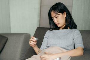Sick asian woman reading clinical thermometer while cough on a sofa bed at home. photo