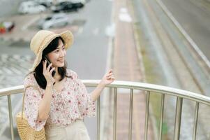 young asian woman traveler with weaving basket using mobile phone and standing on overpass with railway background. Journey trip lifestyle, world travel explorer or Asia summer tourism concept. photo