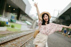 joven asiático mujer viajero con Costura cesta contento sonriente mirando a un cámara junto a tren ferrocarril. viaje viaje estilo de vida, mundo viaje explorador o Asia verano turismo concepto. foto