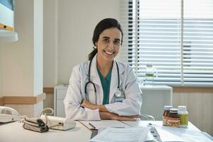 Portrait of beautiful female doctor and professional nutritionist in uniform arms crossed and smiling with supplementary foods on desk for healthy diet at hospital. photo