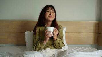 Beautiful young Asian woman wearing sweatshirt smiling and drinking tea on bed in white apartment video