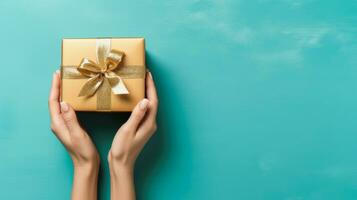 Top view of hands holding gold gift box with copy space on turquoise background, Generative ai. photo