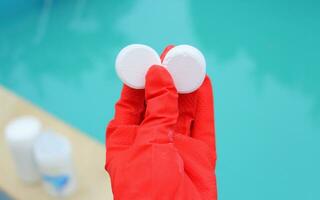 White small chlorine tablet in the hand of a red protective glove of a pool disinfection service worker. The beginning of the swimming season in the hot summer on vacation. photo