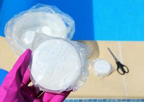 A white large chlorine tablet in the hand of a purple protective glove of a pool disinfection service worker. The beginning of the swimming season in the hot summer on vacation. photo