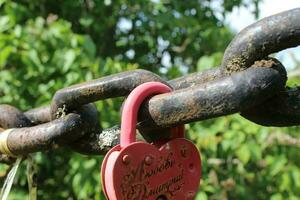 Red love padlock attached to chain. Rusty lock with heart shape carved hanging outdoors. Valentines day, unity, memory concepts photo