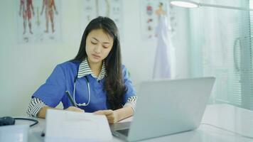 giovane femmina infermiera scrittura Appunti utilizzando il computer portatile a posto di lavoro scrivania. donna professionale medico indossare bianca cappotto Lavorando su computer controllo appuntamenti, fare ricerca o rapporto nel ospedale ufficio. video