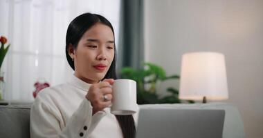 metraggio di contento giovane asiatico donna utilizzando un' il computer portatile computer mentre dire bugie su il divano nel il vivente camera. benessere a casa, rilassante e stile di vita concetti. video