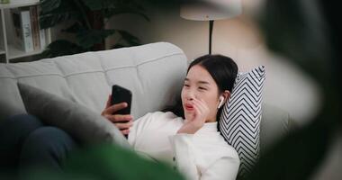 Footage of Happy young Asian woman video call with friends via a smartphone while lying on the sofa in the living room. Wellness at home, relaxing and lifestyle concepts.