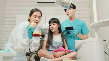 Asian female pediatric dentist and assistant demonstrate toothbrush to little girl with teeth model in dental clinic, well-being hygiene, and professional orthodontic healthcare work in kid hospital. video