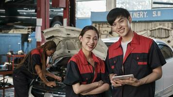 dos asiático profesional mecánica, masculino ingeniero y compañero sonrisa y Mira a cámara, inspeccionar reparar trabajo Lista de Verificación a cochera, Servicio coche mantenimiento, y fijación especialista ocupaciones auto industria. foto