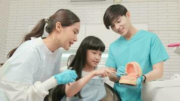 Asian female pediatric dentist and assistant demonstrate toothbrush to little girl with teeth model in dental clinic, well-being hygiene, and professional orthodontic healthcare work in kid hospital. video