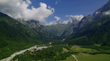 Panoramic aerial view of summer mountain valley landscape with green forest, snowy peaks, mountain river and sky with clouds video