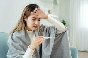 joven asiático mujer teniendo alto fiebre mientras comprobación cuerpo temperatura, hembra estornudos y líquido nariz con estacional influenza, alérgico, digital termómetro, virus, coronavirus, enfermedad, respiratorio foto