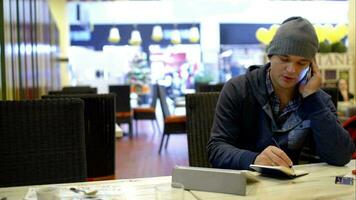 jovem homem dentro cafeteria fazer notas enquanto falando em telefone video