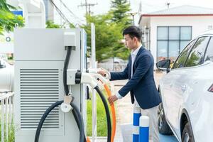 Asian man with EV Car or Electric vehicle at charging station with the power cable supply plugged on electric car charging station. Concept of green energy and reduce CO2 emission. photo