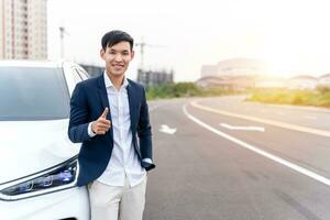Asian man looks on camera while standing near new electric car with thumbs up on city street. Electric car. EV Car. EV vehicle. Concept of green energy and reduce CO2 emission. photo