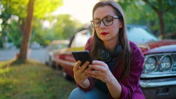 Woman is crouched down next the red vintage car and using smartphone video