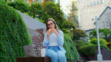 mulher tendo uma almoço com uma sanduíche e café dentro a parque com uma computador portátil video