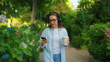 donna con caffè e smartphone a piedi attraverso il fioritura giardino video