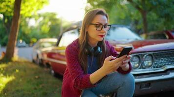 Woman is standing crouched down next the red vintage car and using smartphone video