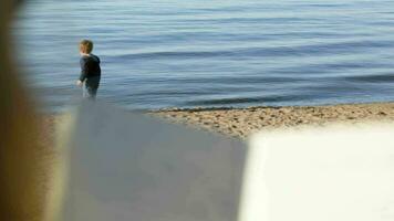 Boy at the water playing with wooden stick, mother reading video