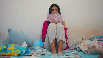 an Asian woman with black hair holding a lot of money with a very sad expression in her room video