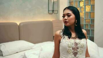 un asiático mujer en un blanco vestir se sienta en un cama y mira sarcásticamente a su novio mientras en un habitación video
