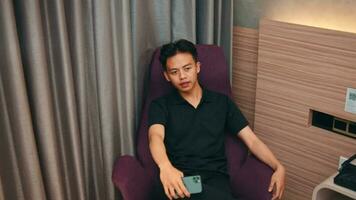 un asiático hombre es sentado en un púrpura sofá mientras esperando para su Novia en un hotel habitación video