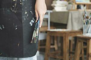 A close-up of the artist hands wearing an apron smeared with paint. Clutching many brushes and paintbrushes. photo