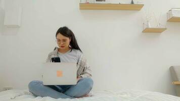 Asian  female wearing eyeglasses messaging with smartphone and a notebook laptop computer on bed at home. photo