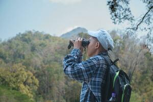 Asian boy in plaid shirt and cap doing the summer activity at local national park by watching birds, fish, insects, animals, trees, flower by using map and binoculars, soft and selective focus. photo