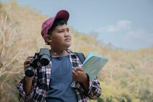 Asian boy in plaid shirt and cap doing the summer activity at local national park by watching birds, fish, insects, animals, trees, flower by using map and binoculars, soft and selective focus. photo