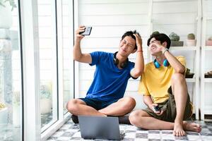 Young asian gay couple spending their free time, making selfie and looking after cactus pots on shelves behind by using magnifying glass, LGBT lover special recreational activity concept. photo