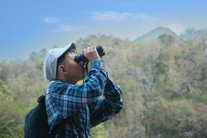 Asian boy in plaid shirt and cap doing the summer activity at local national park by watching birds, fish, insects, animals, trees, flower by using map and binoculars, soft and selective focus. photo