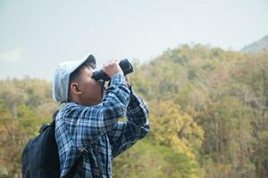 Asian boy in plaid shirt and cap doing the summer activity at local national park by watching birds, fish, insects, animals, trees, flower by using map and binoculars, soft and selective focus. photo