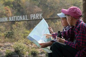 asian boys holds binoculars and local national park map, resting and reading information in map near sling bridge of local national park during nature and summer camp of them, teens' activity concept. photo